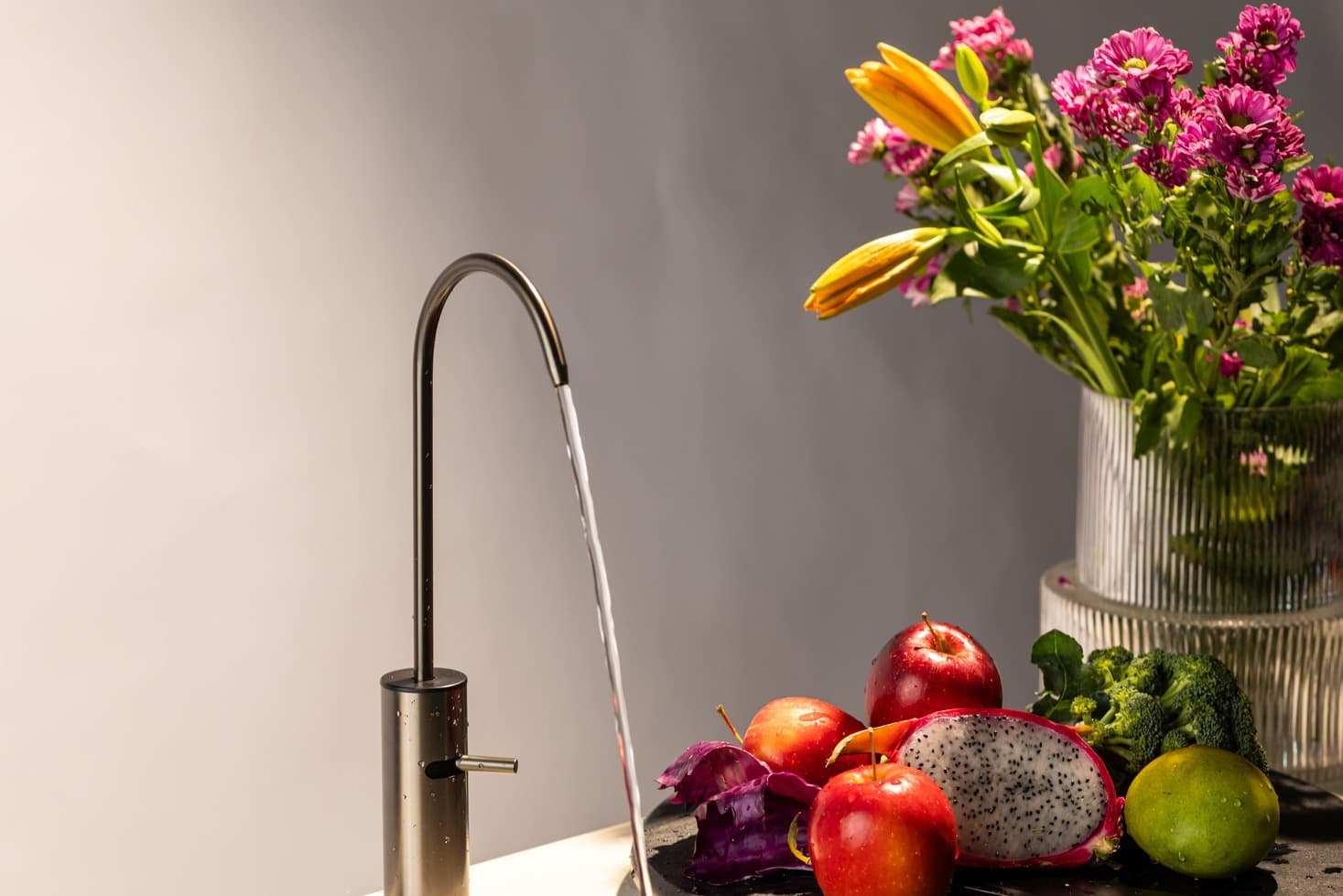 Water pouring from a Boon tap into a black sink beside fresh flowers and fruit in warm light