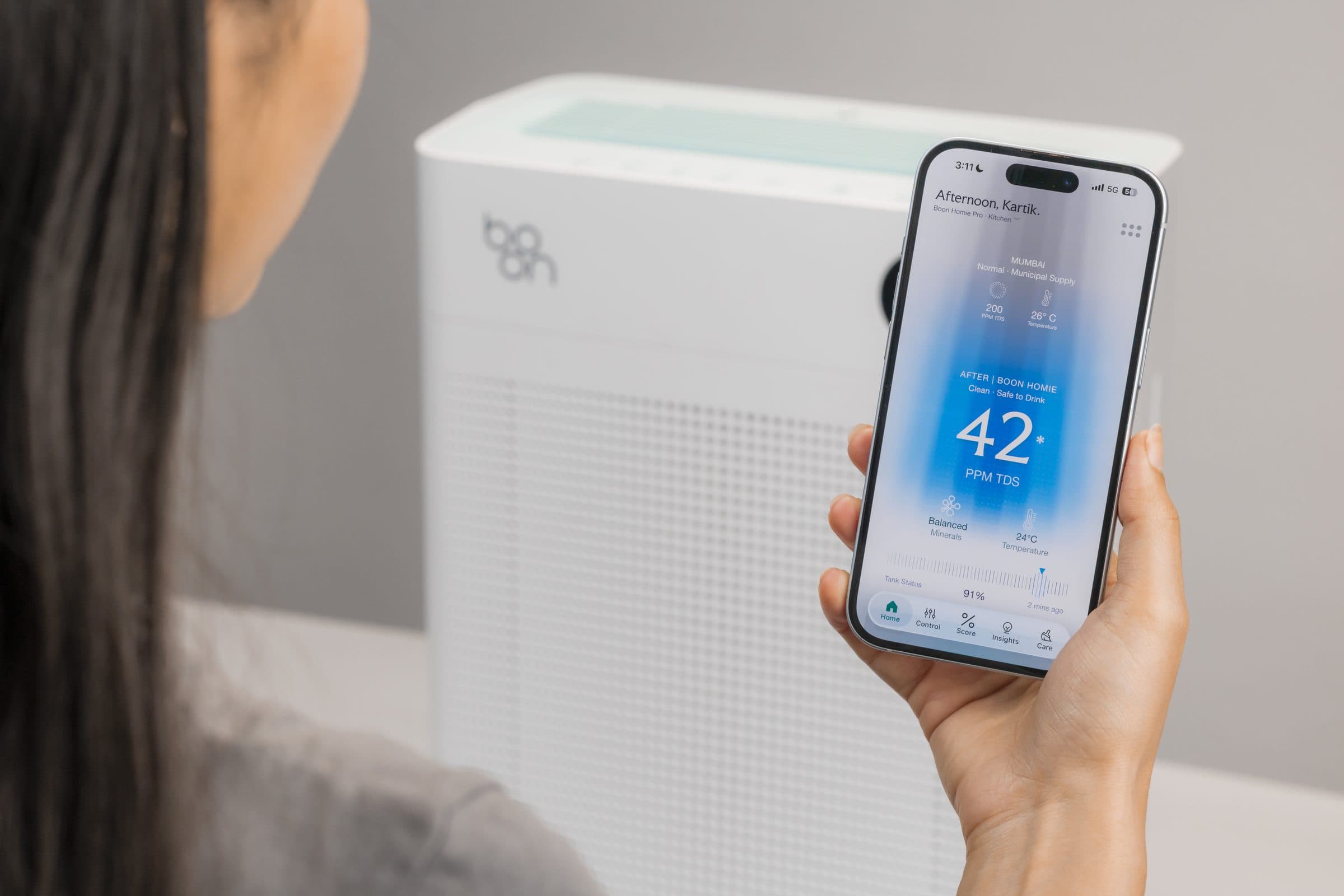 A hand holding the Boon app up in front of Boon Airvana™: Mumbai's outdoor AQI of 356, the room's AQI of 39 after Airvana™, and the same 39 on the unit's display