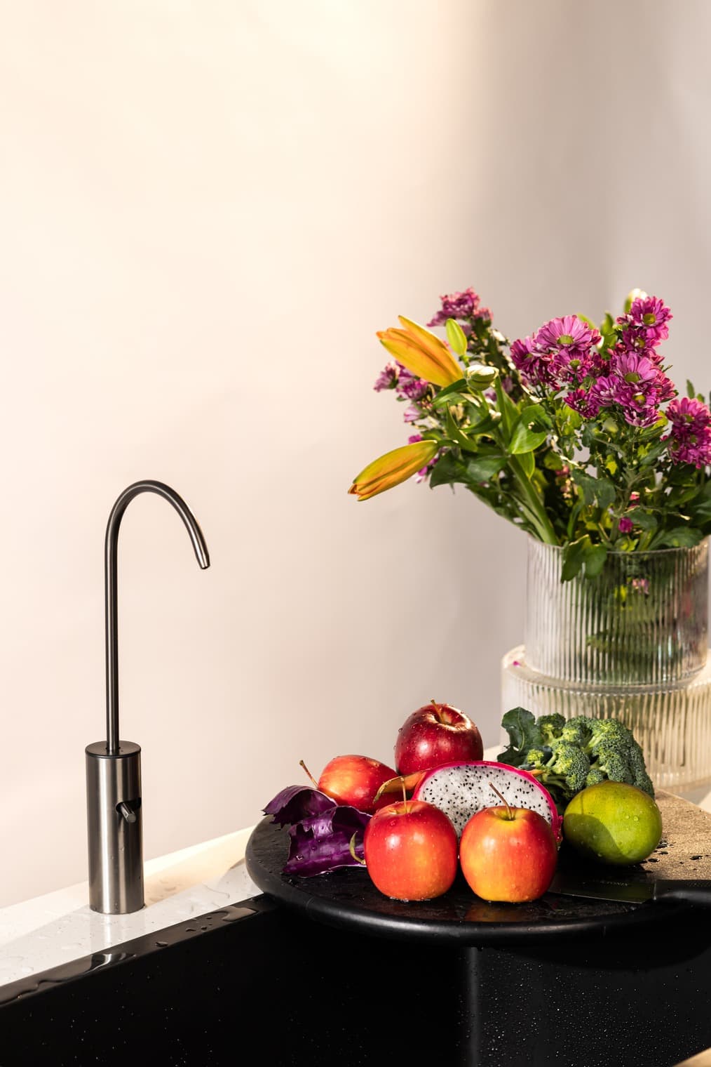 A Boon tap on a kitchen counter beside fresh produce and flowers.