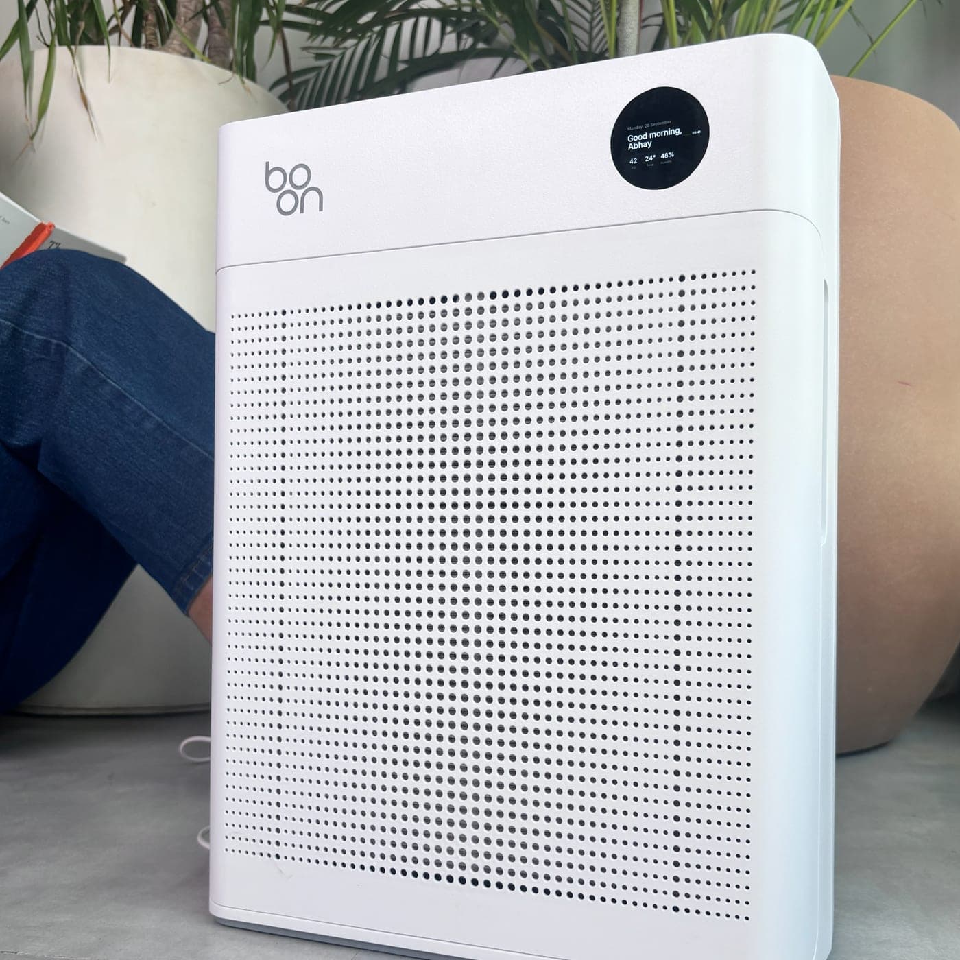 Boon Airvana™ in a living room beside potted plants, its display showing a morning greeting